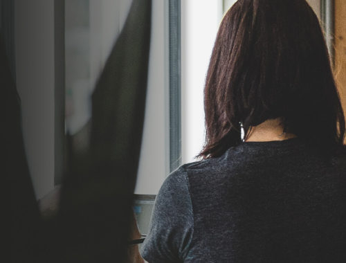 Woman with shoulder length hair in a dark gray shirt looking out a window
