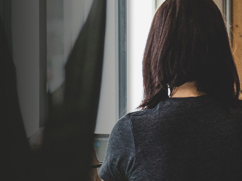 Woman with shoulder length hair in a dark gray shirt looking out a window